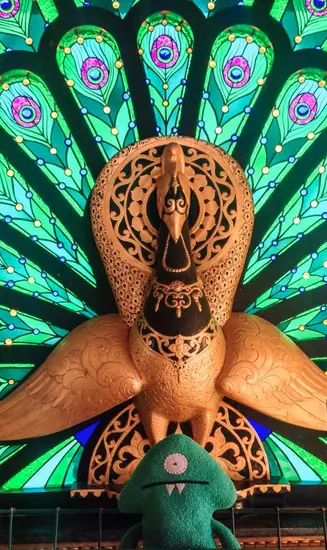 Keltar seated before a carved orange peacock with a fan of green stained-glass feathers