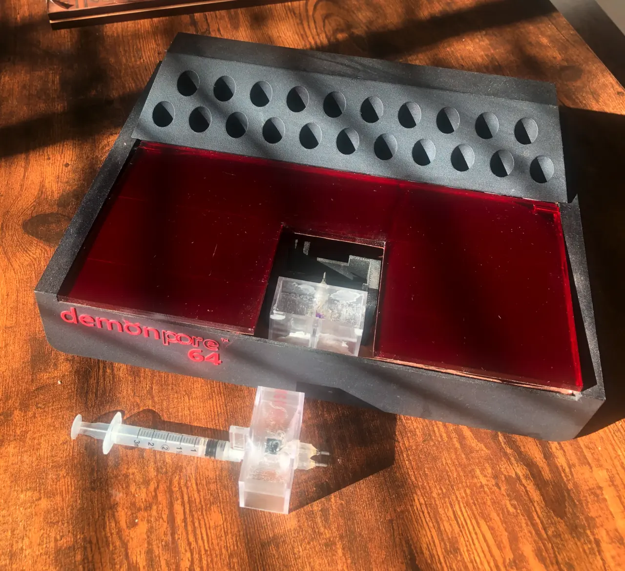 Matte black demonpore 64 cartridge tray on a wood table: two rows of teardrop-shaped wells along the back, a red translucent inset with a clear flow cell in the centre, a plastic syringe in front, red lettering demonpore 64 on the front edg