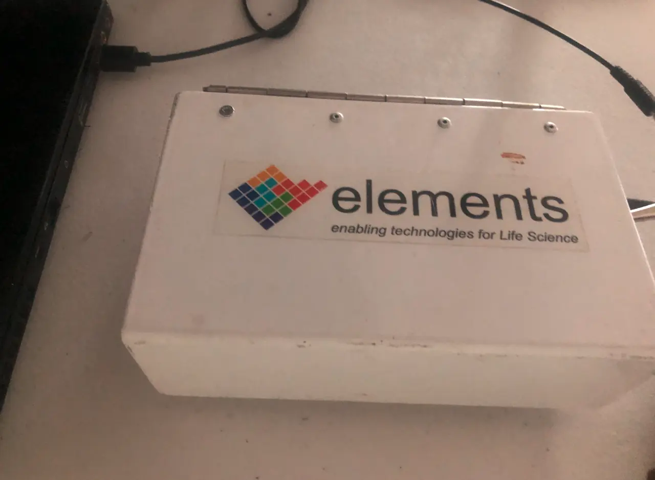 White hinged Faraday-style enclosure carrying a third-party label (elements, enabling technologies for life science) with cables trailing out.