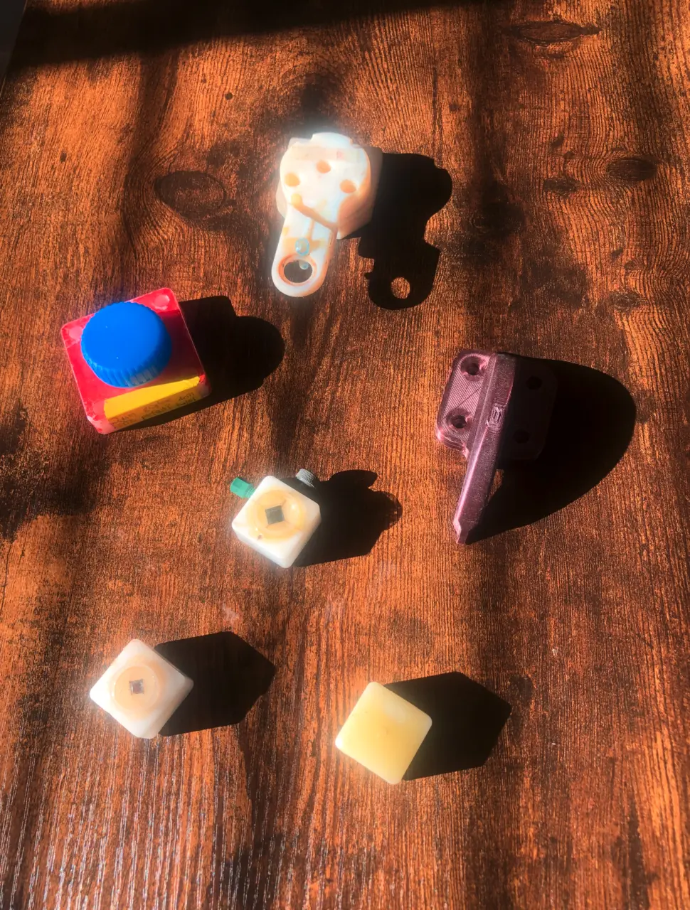 Small square 3D-printed parts in white, yellow and orange on a wooden table, with a purple pusher and a red block