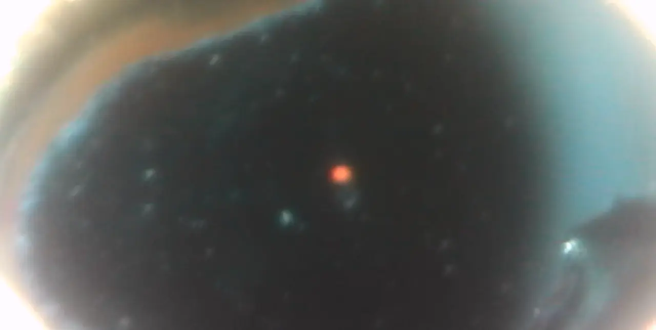 Dark-field microscope view of a laser-drilled pore: a dark oval opening with a single red laser dot and floating specks (1334x672).